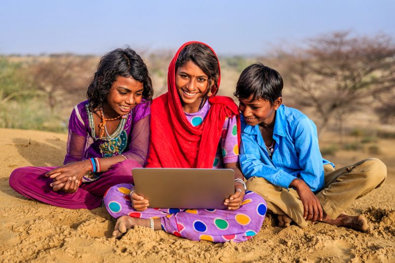 Happy Indian children, sitting on a sand dune and using laptop in desert village, Thar Desert, Rajasthan, India.