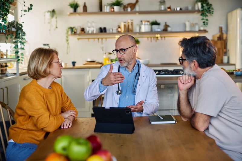 A male doctor talks with a senior couple in their cozy kitchen, using a tablet to explain health information and treatment options during a calm, personal home healthcare consultation.