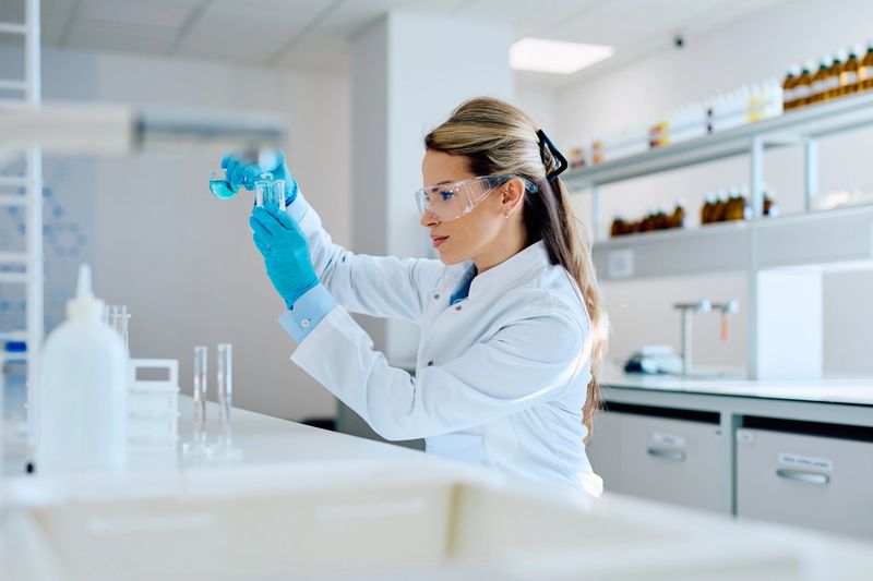 Young woman scientist in protective eyewear and gloves pouring blue liquid into a test tube during chemistry research