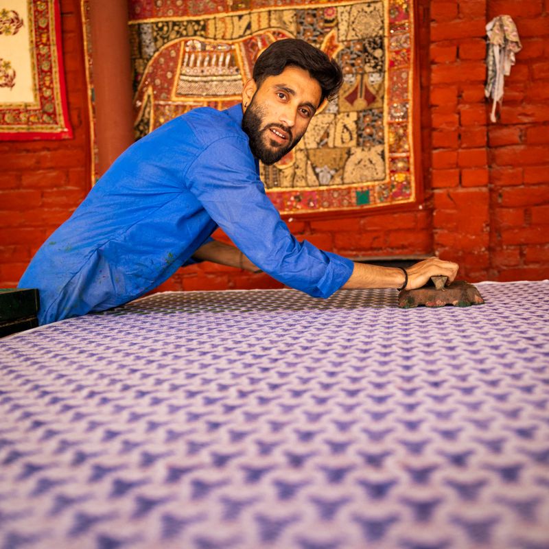 A craftsman in a bright blue shirt leans over a large piece of patterned fabric, pressing a wooden block to create a repeating purple motif. He glances toward the camera while working in a workshop with a red brick wall and decorative embroidered textiles hanging behind him. The image captures traditional hand-block printing and focused, manual textile craftsmanship.