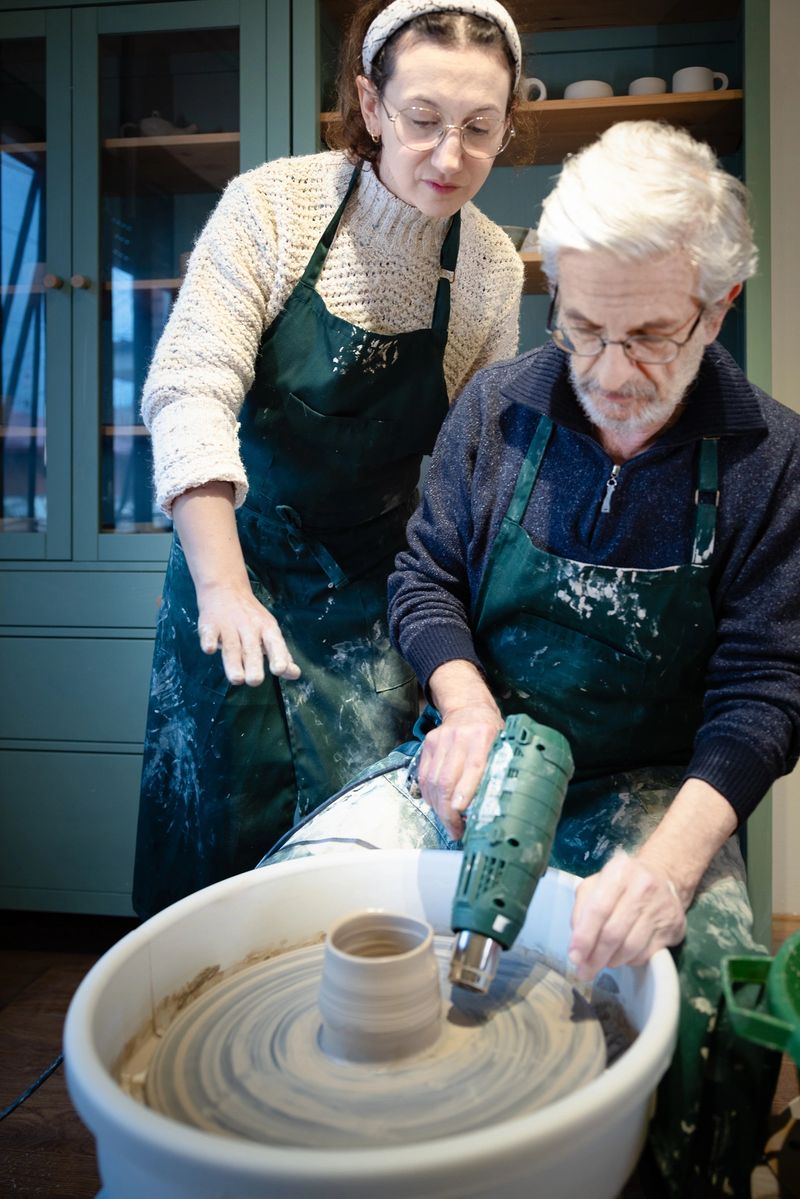 A woman pottery teacher instructs an elderly male student how to dry a newly made wet ceramic cylinder with a heat gun the pottery wheel, while completed ceramics pieces decorate the shelves of the studio in the background.