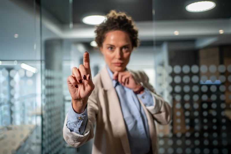 Confident businesswoman in a glass office interacting with a futuristic virtual touchscreen, pointing to select data and demonstrating technology-driven leadership and innovation