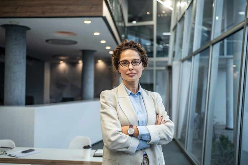 Confident mid adult businesswoman standing with arms crossed, wearing glasses and a blazer, representing leadership, success, and professional expertise in a contemporary office environment
