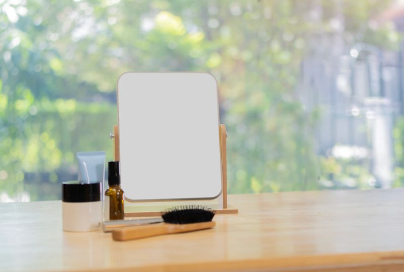 Tabletop makeup mirror, Mirror, cosmetic products and wooden comb on wooden table in a glass-walled room with background of nature outside.