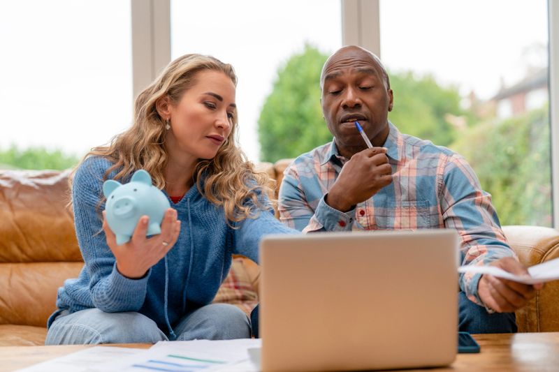 Two adults work on their finances at home. One person holds a piggy bank while the other looks at a laptop. Papers are spread out on the table around them.