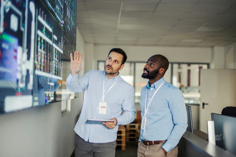 Two adult male engineers, a mid adult and a young adult, collaborating and discussing crucial data visualization on a large digital screen in a modern control room