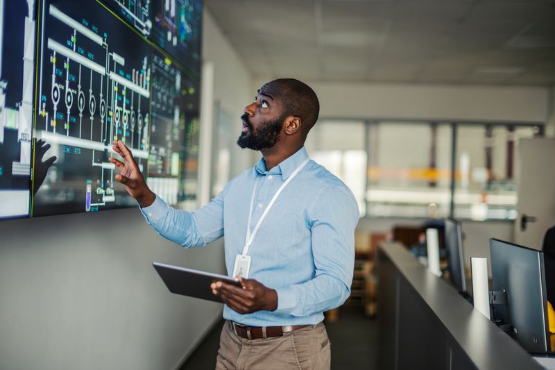 African american engineer holding a digital tablet and observing complex data on a large screen, managing industrial operations in a modern control room