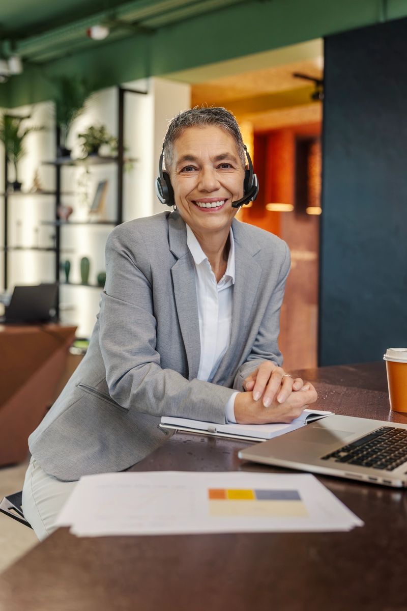 Mature adult businesswoman smiling while looking at camera and wearing a headset, providing customer support in a modern office environment with a laptop and documents on the table