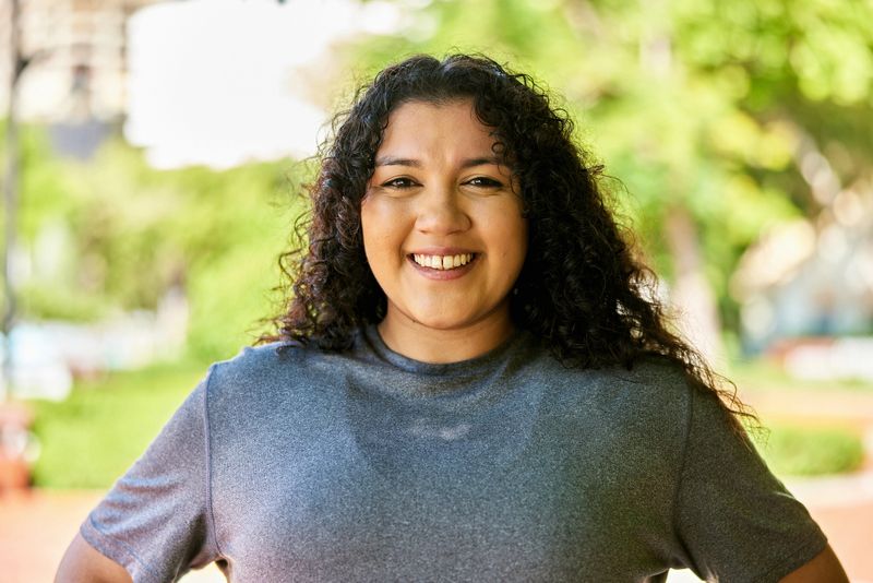 Head and shoulders view of casually dressed individual with long curly hair, hands on hips, smiling at camera.