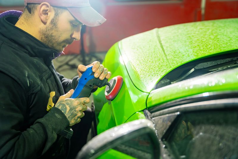Young man polishing a vibrant green car with an orbital polisher in a professional detailing garage, concentrating on paint correction to achieve a flawless high-gloss finish