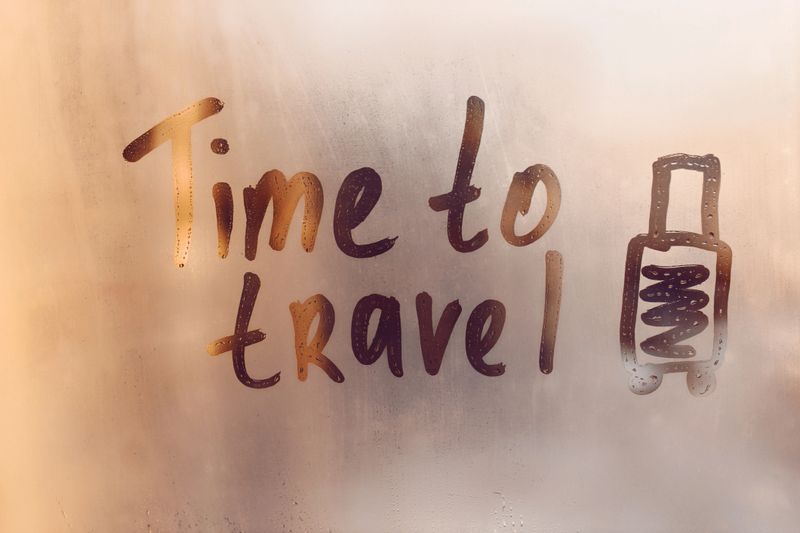 Handwritten message Time to travel and doddle shape of suitcase on glass of sunset window with raindrops