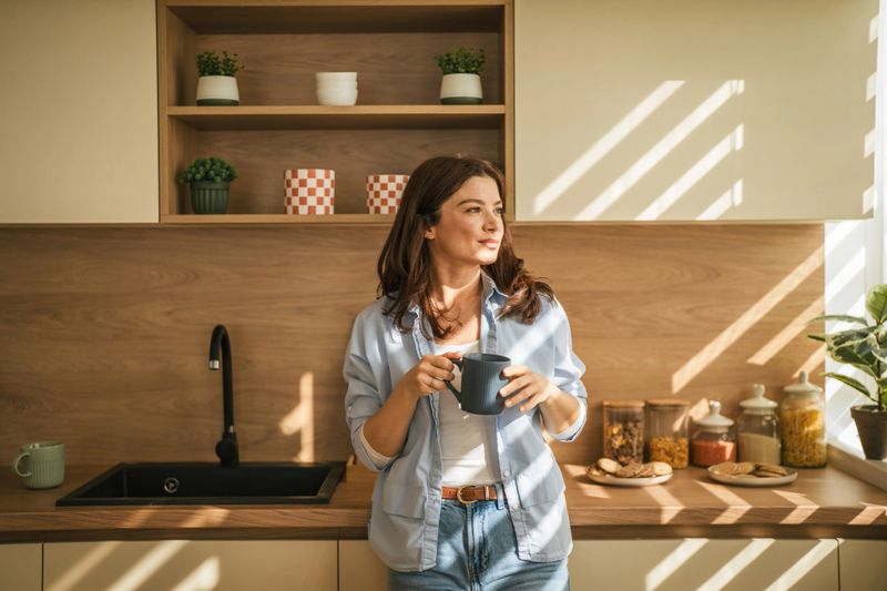 Woman standing in a modern kitchen holding a mug, illuminated by natural sunlight stripes, looking thoughtfully into the distance and enjoying a quiet relaxed moment