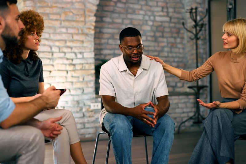Man sitting in a circle shares his thoughts while other participants listen and offer support during a group therapy meeting.