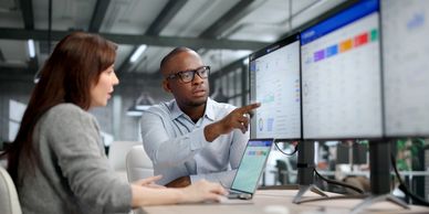 Two professionals discussing data on dual monitors.