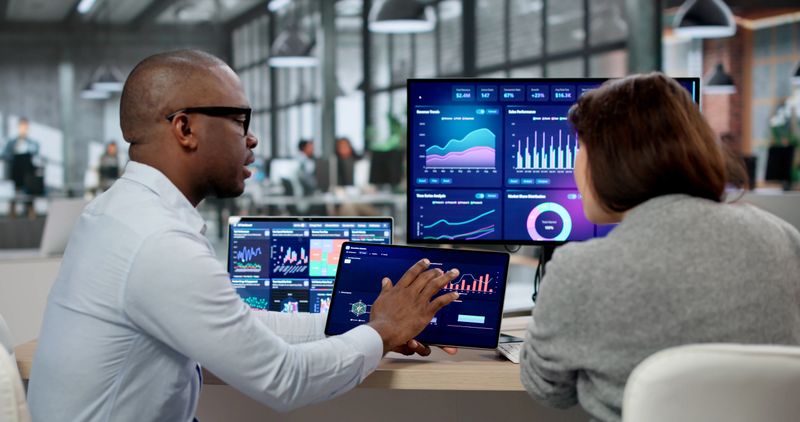 Business Analyst And Marketing Coach Examine KPI Dashboards On Multiple Screens. An African American Woman