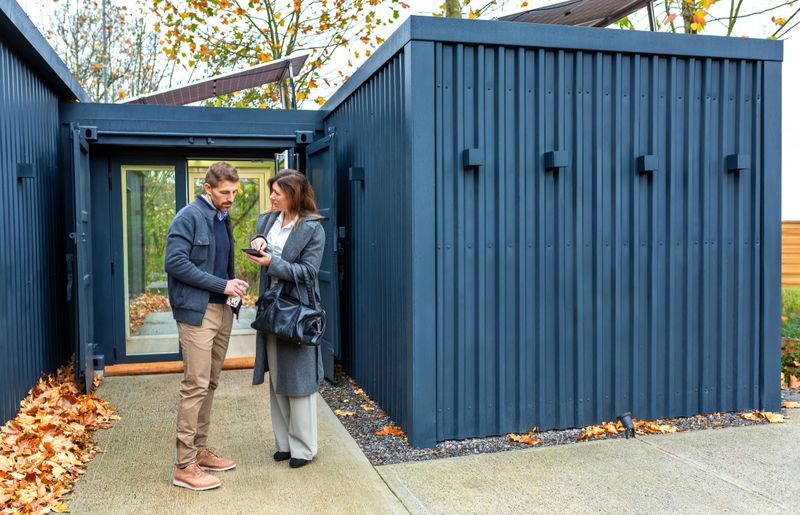 Architect explaining design concepts of modern modular house built from repurposed shipping containers with solar panels in mobile phone to a satisfied client, with autumn leaves on the ground