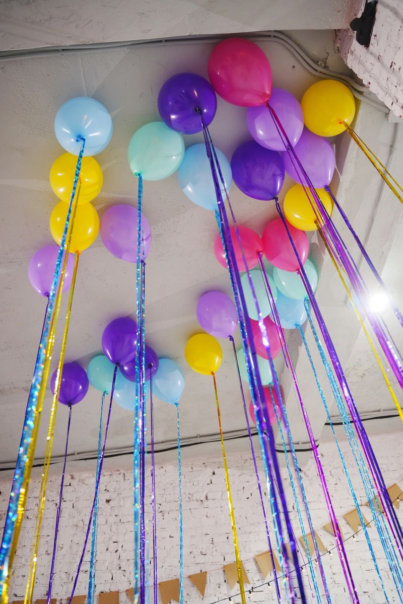 Brightly colored balloons with long strings are suspended from the ceiling. The scene shows a party atmosphere with various colors creating a fun mood.