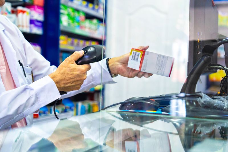Close-up of a pharmacist in white coat scanning a medication box with a barcode reader at a pharmacy counter. Drug dispensing, retail healthcare, prescription medicine, and point of sale concept.