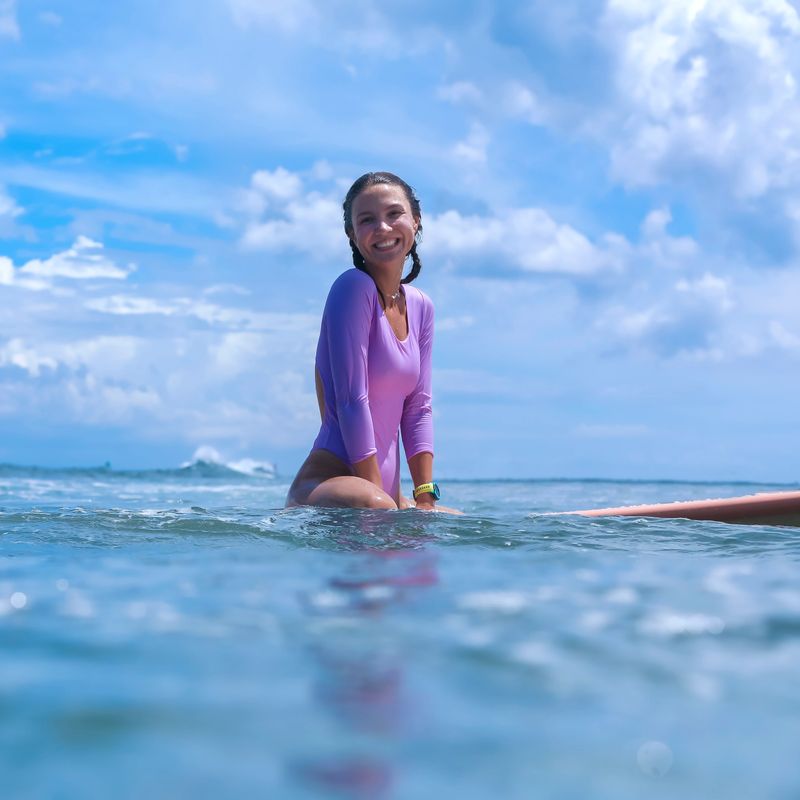 Young Woman Surfer Sitting On Board Smiling Waiting Amid Calm Ocean And Blue Sky. Purple Rashguard, Gentle