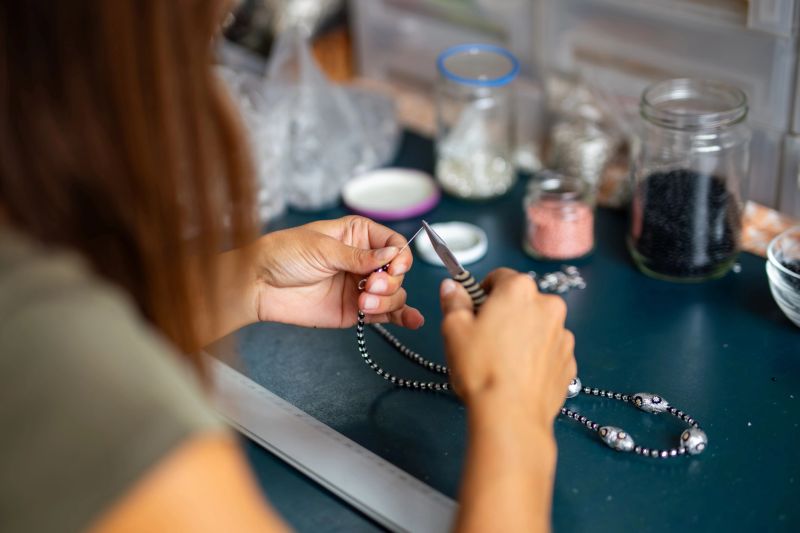 A female artisan is crafting a necklace at a workbench. She uses pliers to manipulate a strand of beads, indicating detailed jewelry work. Tools and materials, including jars filled with various beads, are organized on the table. The setting suggests a professional workspace for handmade jewelry creation, reflecting the meticulous process involved in goldsmithing.