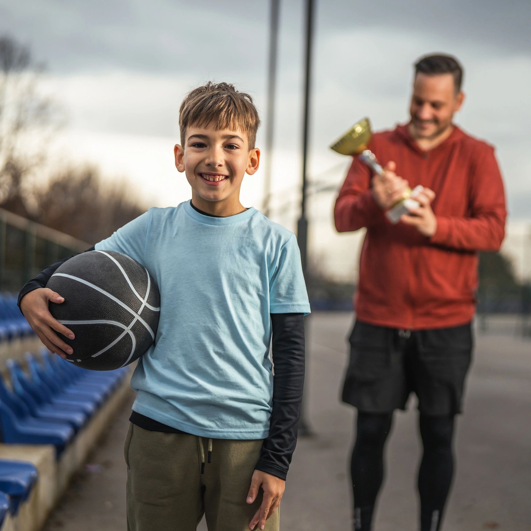 Smiling boy holding basketball with man holding trophy in background.