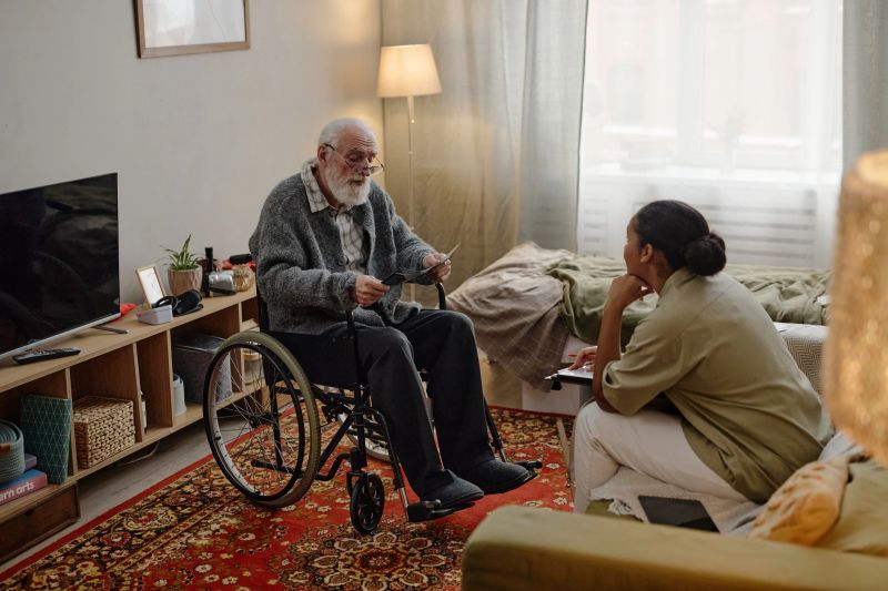 Senior Caucasian man with disability sitting in wheelchair, holding photographs while young adult Black woman social worker listening attentively and taking notes in residential room