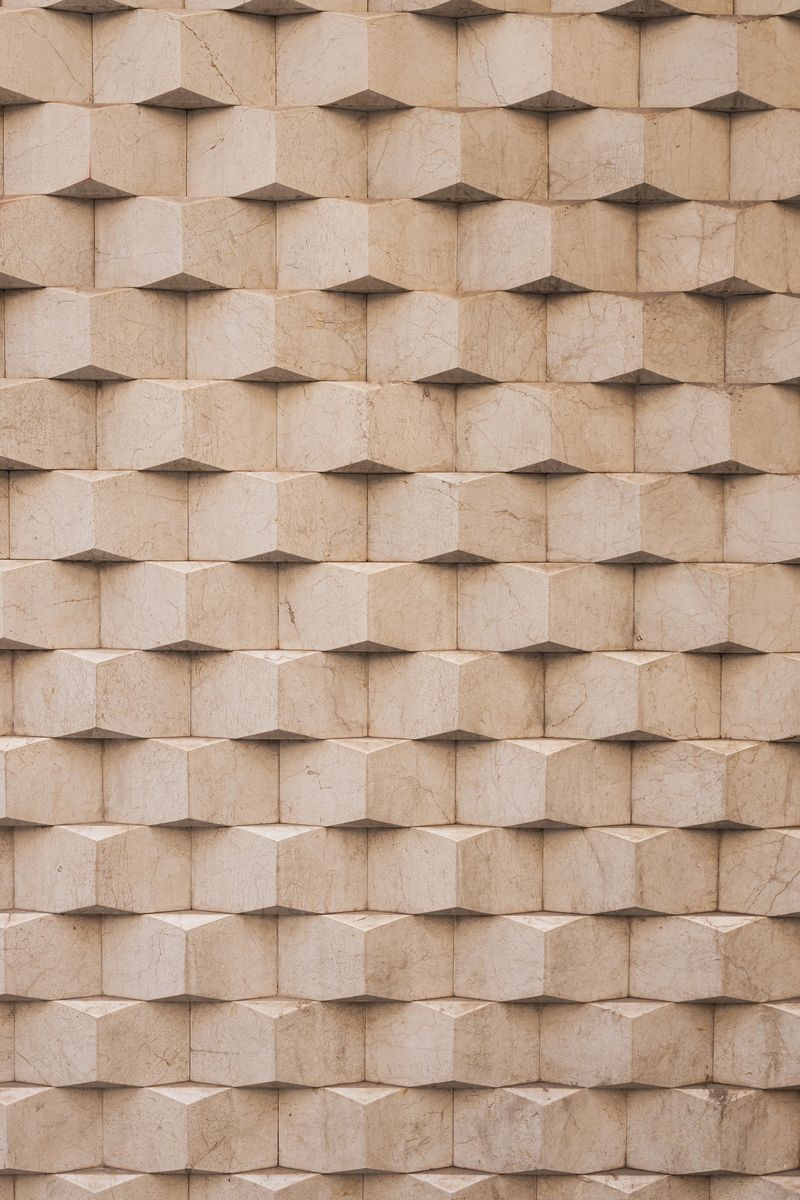 Beige marble tiles forming a repeating geometric pattern on an architectural facade, creating a textured surface with an optical illusion of depth and shadow