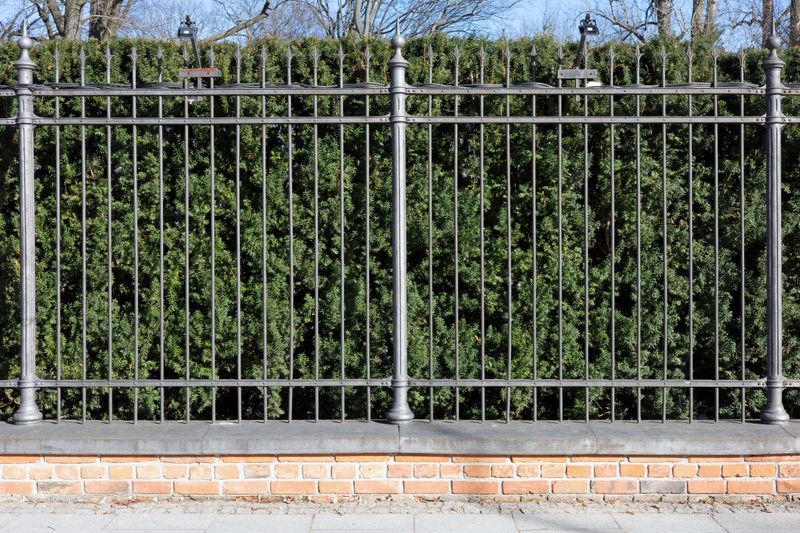 This high-quality architectural photograph features a meticulously crafted grey metal fence mounted on a sturdy brick and stone foundation. The central focus is a prominent, ornate decorative pillar with a spherical and pointed tip, dividing the symmetrical sections of the vertical iron bars. Each bar is topped with a sharp, spear-like finial, adding both aesthetic appeal and a sense of security.The background is entirely filled with a lush, vibrant green coniferous hedge, likely yew or cedar, which provides a natural, soft contrast to the rigid, cold texture of the ironwork. The base of the structure consists of a weathered red brick wall topped with a smooth, grey stone coping. Bright, direct sunlight casts subtle shadows, highlighting the textures of the metal, the rough brickwork, and the intricate needles of the foliage. Two small floodlights are visible at the very top corners, suggesting the area is illuminated at night. This image perfectly captures themes of privacy, security, classic landscaping, and high-end property boundaries. The composition is clean, centered, and balanced, making it ideal for architectural, real estate, or home improvement editorial content.