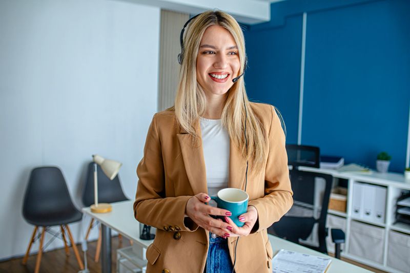 Smiling professional woman wearing a headset and holding a mug while speaking with clients during a comfortable and flexible work routine.