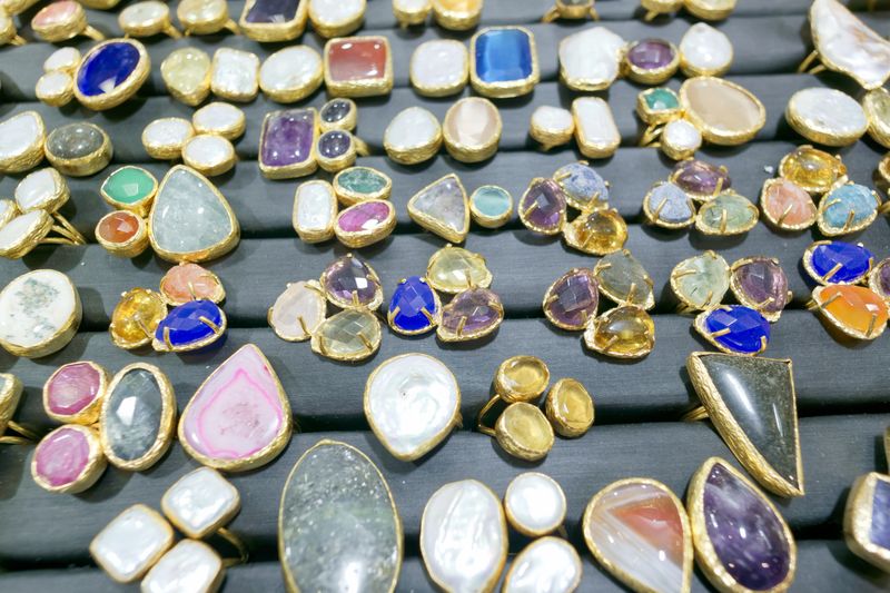 Many rings with colorful gems are arranged on a table in a market. The rings have different shapes and sizes, showing a variety of stones. The setting is bright and lively.