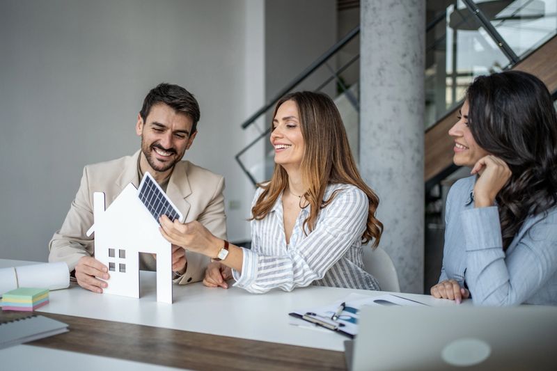 Group of smiling business people collaborating on a sustainable home project, examining a model house with a solar panel during a meeting in a modern office