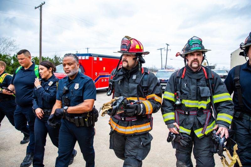 Group of firefighters and emergency personnel assembled outdoors at fire station. Multiple responders wearing protective gear including helmets and turnout gear stand together near red fire truck. Team includes male and female firefighters of diverse backgrounds. Clear daytime conditions with utility poles and station buildings visible in background. Scene captures active emergency services personnel during operational coordination.