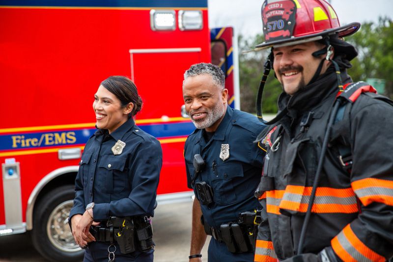 Three emergency responders stand together outside a fire station in front of a red fire engine. A female Hispanic paramedic in dark blue uniform stands on the left, a male Black paramedic in dark blue uniform stands in the center, and a male firefighter wearing a red helmet and dark gear with orange reflective stripes stands on the right. All three are smiling and appear relaxed during their shift at the station.