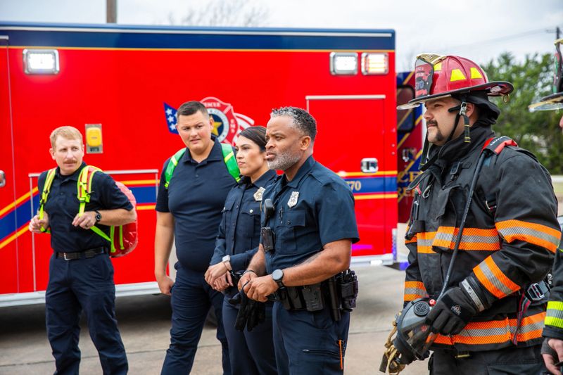 Group of emergency responders gathered outside fire station next to red fire truck. Team includes firefighters wearing protective gear with helmets and reflective stripes, and paramedics in dark blue uniforms. One firefighter on right wears full protective equipment with orange reflective gear and helmet. Officers appear to be in discussion or briefing. Red fire engine visible in background. Daytime outdoor setting at emergency services facility.