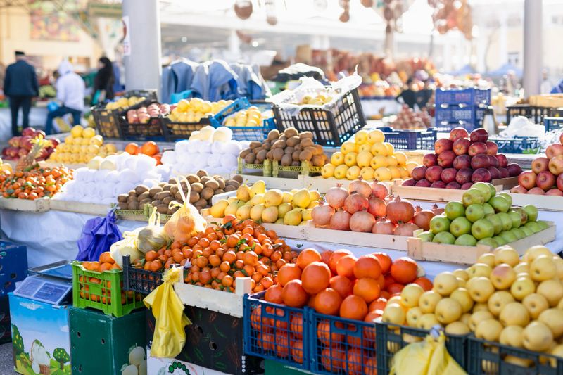 Sunny farmer's  market stall display abundant seasonal fruits. Pyramids of juicy oranges, mandarins, apples in crates, baskets. Lively oriental trading atmosphere. Samarkand Siab market, Uzbekistan.