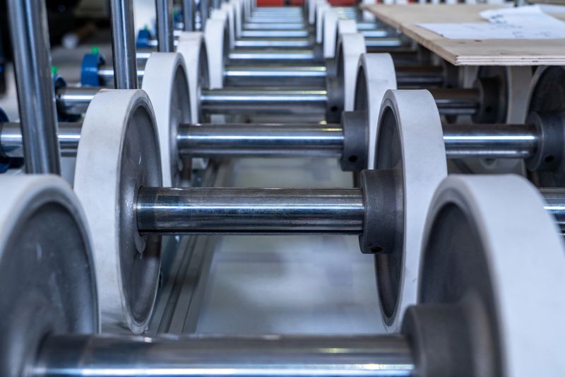 A professional close-up shot of an industrial roller conveyor system. The image features a perspective view of multiple stainless steel shafts fitted with large white rubber-coated wheels, designed for the smooth transport of heavy materials.