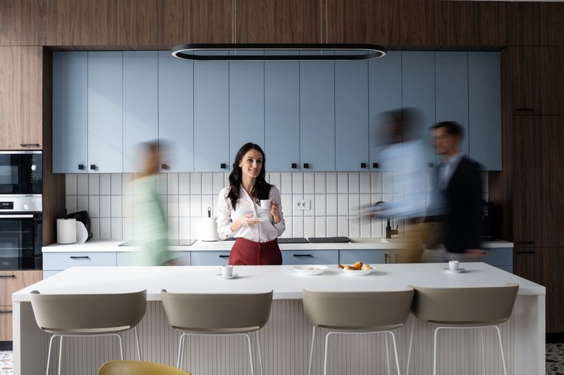 Young businesswoman smiling, holding a cup of coffee, in a dynamic coworking kitchen while colleagues with motion blur passing by