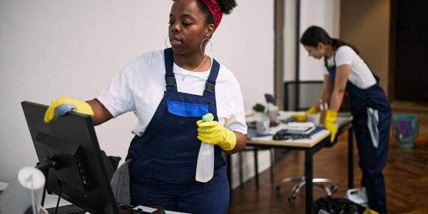 Two women cleaning an office, wearing gloves and overalls.