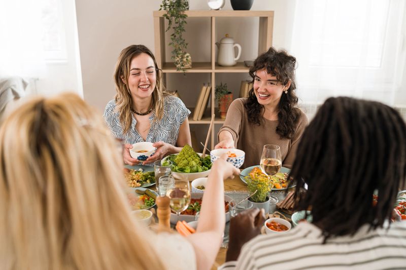 A diverse group of young female friends is gathered around a dining table, sharing a meal. They are smiling and engaging in conversation while passing dishes. The table is filled with various colorful dishes and drinks, creating a lively atmosphere.