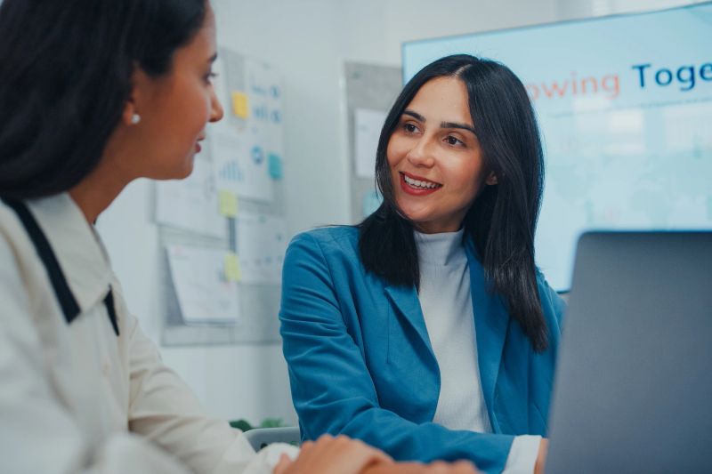 Two professional women collaborate strategy reviews project meeting in office. Reviewing information on laptop while discussing plan and next steps. Teamwork, strategy, and structured collaboration.