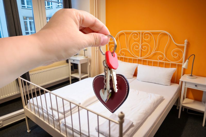 Hand holding keys with keychain in hotel room, apartment, furnished bedroom, vacation rental, bright room, Cozy Beds in Hotel, comfortable stay, new home, romantic getaway, welcoming atmosphere