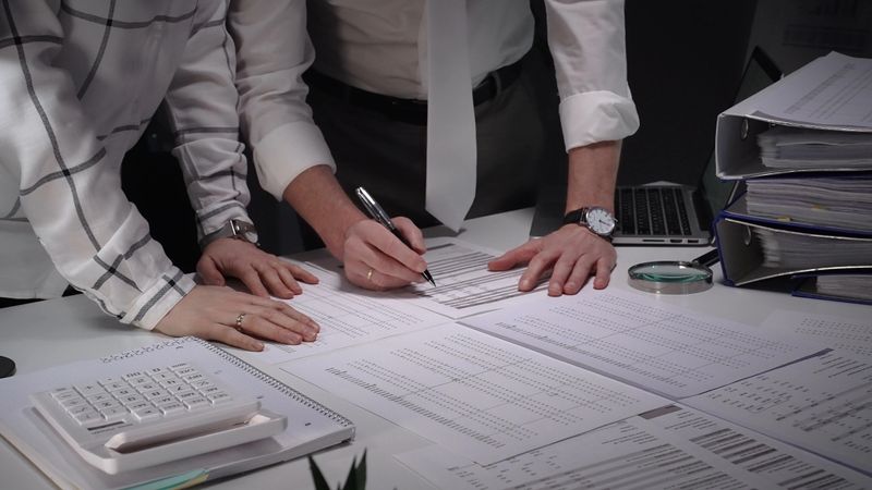 Business professionals at a desk closely examining financial documents with magnifying glass and pen, conducting a meticulous audit or investigation for accuracy and risk assessment