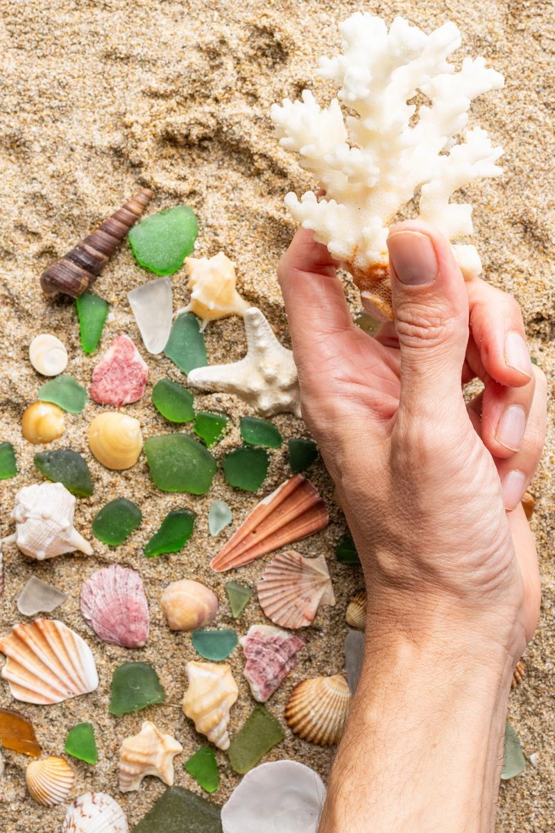 Person's hand gently picking up a piece of white coral from a collection of various seashells and green sea glass spread across light-colored beach sand, indicating a relaxed summer activity