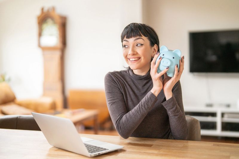 A woman sits at a table holding a piggy bank with a big smile. She looks excited while working on a laptop in a cozy living room during the day.