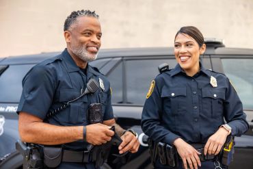 Two police officers smiling and standing together near a patrol car.