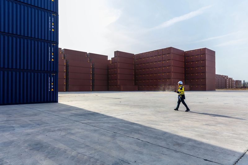 An Asian engineer uses a smart tablet at a dock filled with shipping containers.