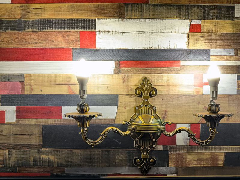 Ornate brass wall sconce with two illuminated bulbs mounted on a colorful reclaimed wooden plank wall, evoking warm, cozy, and vintage interior decor with textured horizontal wood panels and classic detail.