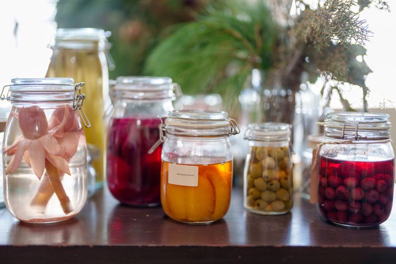A variety of glass jars containing fermented fruits, vegetables, and flowers, including olives, berries, and ginger buds. The colorful jars are displayed on a dark wooden surface in a bright, natural setting with greenery in the background.