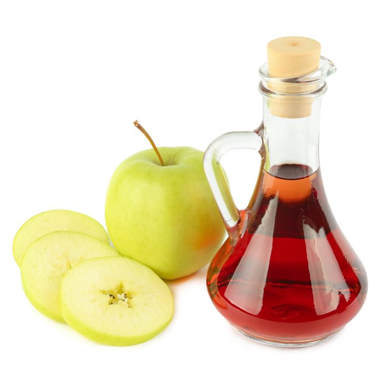 A clear glass bottle filled with red apple cider vinegar stands next to a whole green apple and fresh slices. This clean, minimalist shot on a white background is perfect for health and culinary projects.