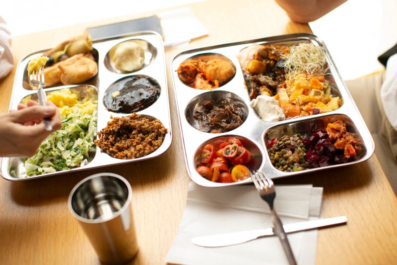 South asian colleagues sharing thali platters in bright office cafeteria with stainless compartmentalized trays, variety